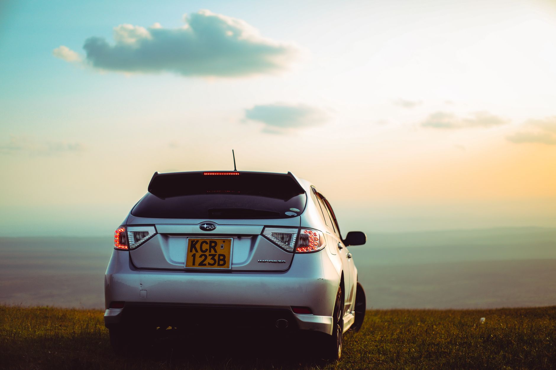 A blue car parked on a dirt road with hills and green trees in the background under a cloudy sky.